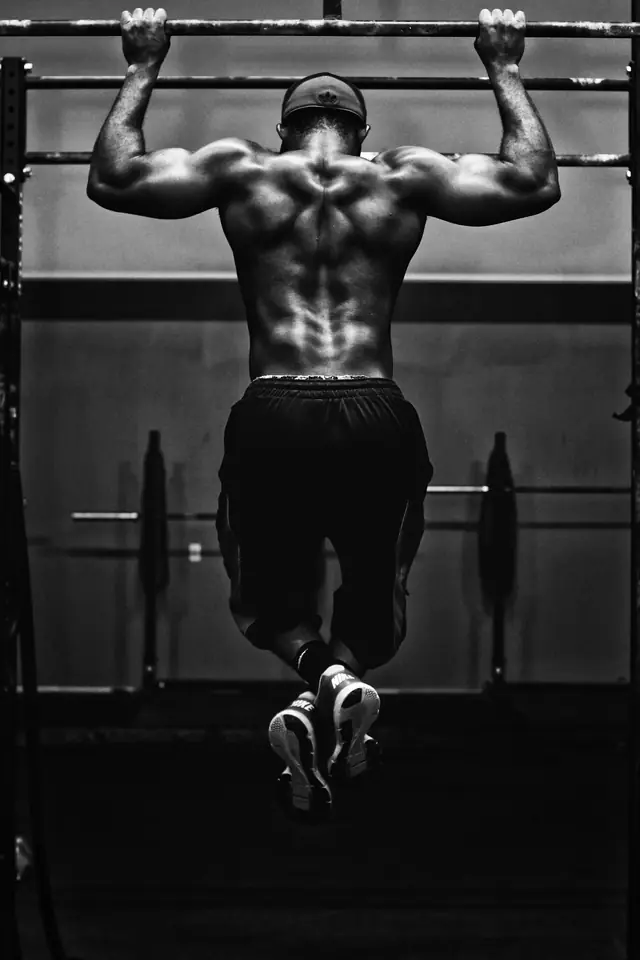 Hombre de espaldas haciendo dominadas en una barra, con la musculatura de la espalda marcada, en blanco y negro