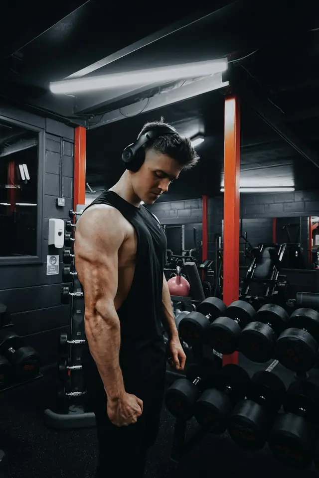 Hombre con auriculares entrenando junto al rack de mancuernas en una sala oscura con columnas rojas